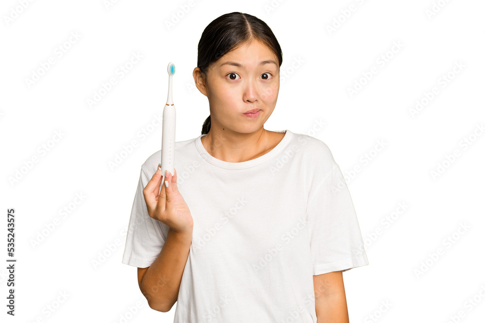 Young Asian woman holding an electric toothbrush isolated on green chroma background shrugs shoulders and open eyes confused. Young Asian woman holding an electric toothbrush isolated on green chroma background shrugs shoulders and open eyes confused.