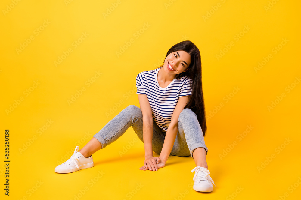 Full length portrait of adorable satisfied malaysian lady sitting floor toothy smile isolated on yellow color background
