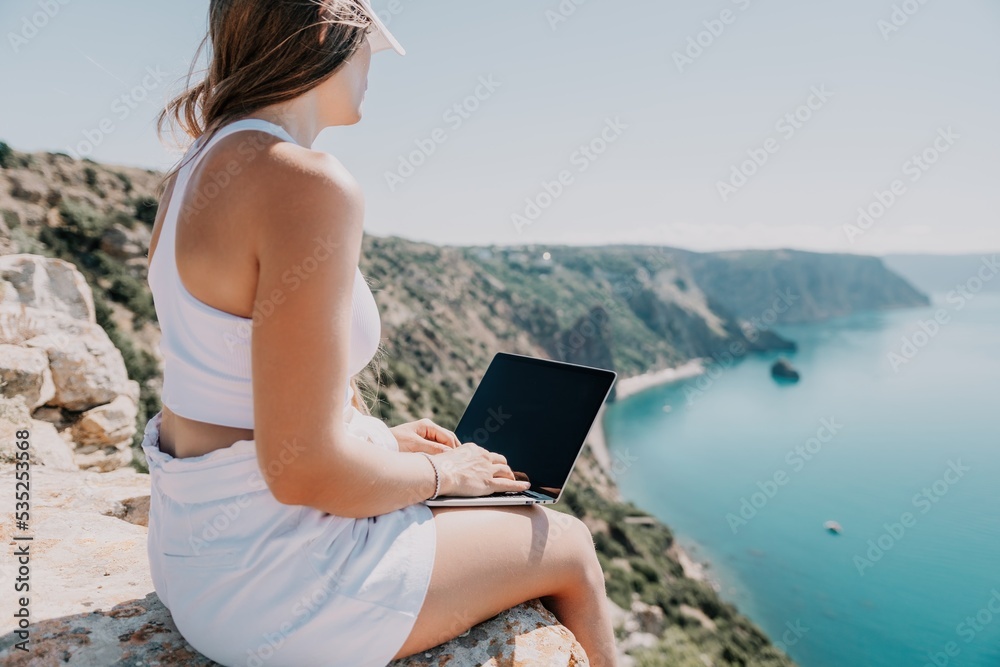 Digital nomad, woman in the hat, a business woman with a laptop sits on the rocks by the sea during sunset, makes a business transaction online from a distance. Freelance, remote work on vacation.