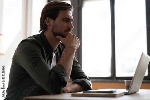 Pensive bearded man sitting at table work at laptop thinking of problem solution