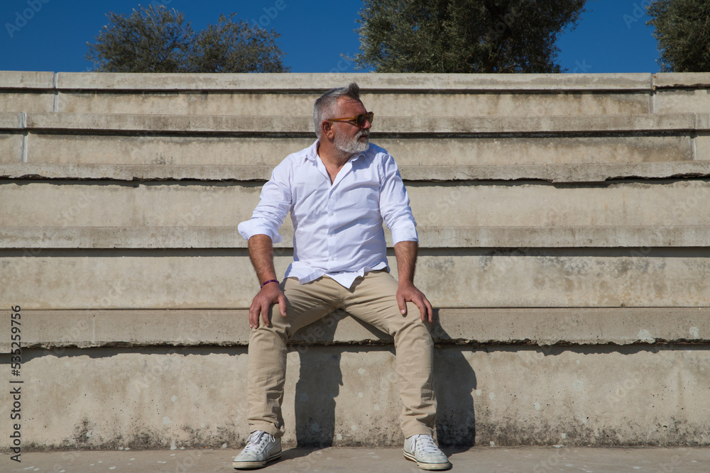 handsome mature man with beard and grey hair is sitting on the steps of an open-air auditorium. The man looks at different places. Senior concept, travel and tourism.