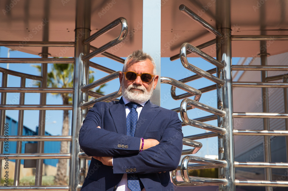 Handsome, mature, grey-haired man with beard, jacket and tie and sunglasses at the entrance to the office building where he works as chief accountant and executive. The man with his arms crossed.
