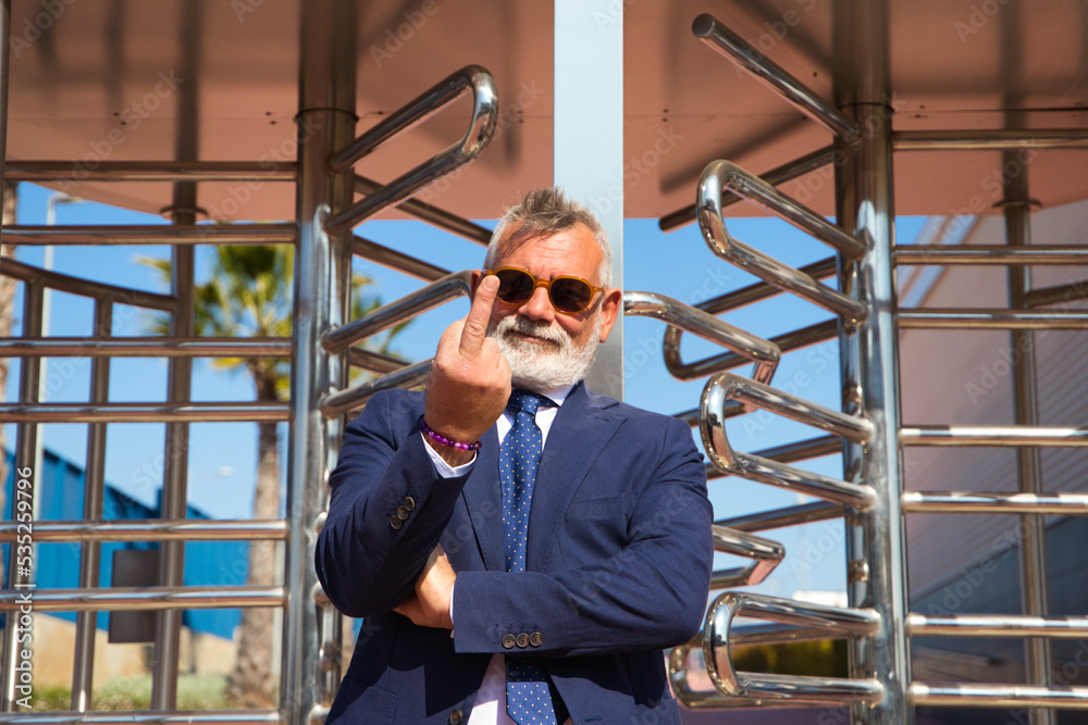 Handsome, mature man with grey hair, beard, jacket and tie and sunglasses at the entrance to the office show Middle Finger or Fuck Signature. Concept of angry or funereal