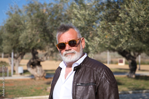 handsome mature man with beard and grey hair is walking in the park. The man is dressed in beige trousers, white shirt and leather jacket. Old age concept