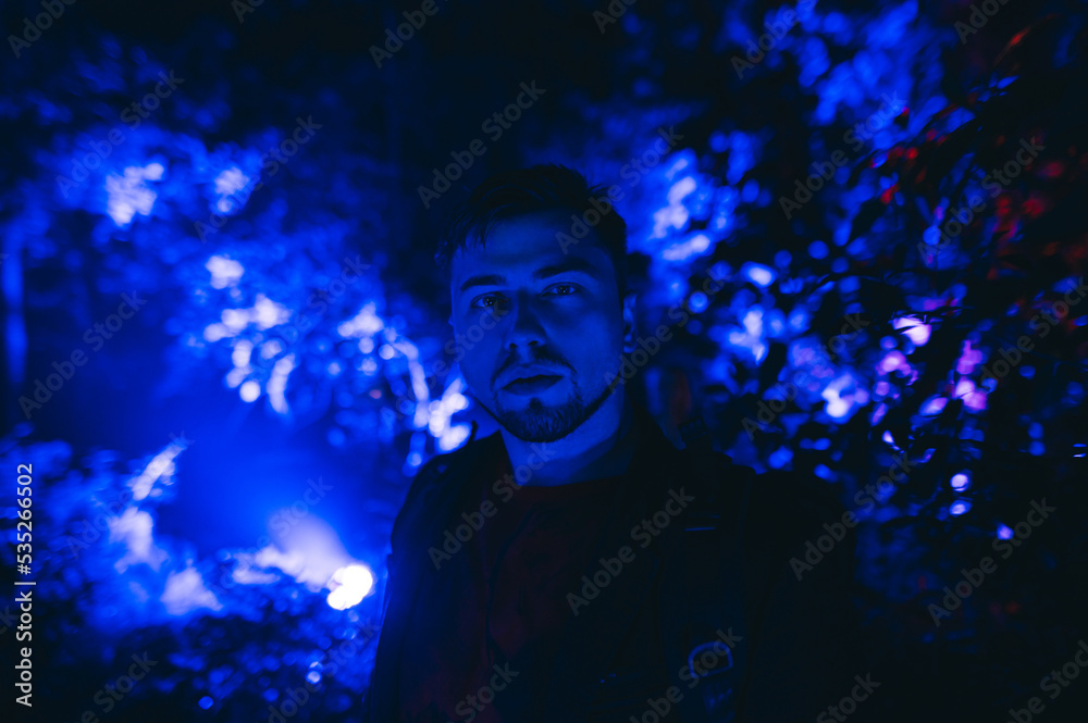 Man with a beard stands at night in the park with blue light on a background of trees and plants