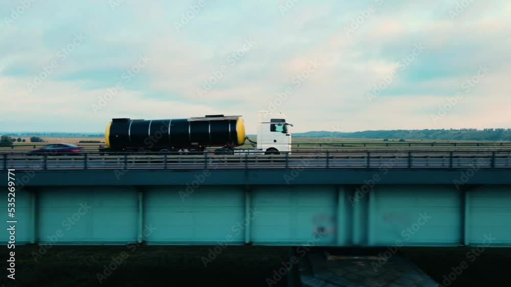 Truck driving with tank trailer on bridge with highway to customer through picturesque rural area with agricultural fields background. Concept of optimizing cargo transportation business Truck driving with tank trailer on bridge with highway to customer through picturesque rural area with agricultural fields background. Concept of optimizing cargo transportation business