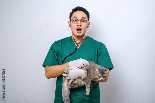 Shocked doctor in vet clinic examining cat with stethoscope, gasping amazed while holding cat in his arm