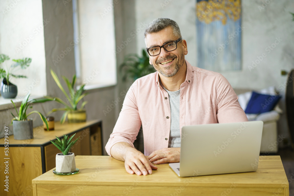 Handsome middle aged man in glasses working sitting at table with laptop at home. Freelancer man working at home using computer. Smiling man using laptop at home. Middle aged businessman at home Handsome middle aged man in glasses working sitting at table with laptop at home. Freelancer man working at home using computer. Smiling man using laptop at home. Middle aged businessman at home