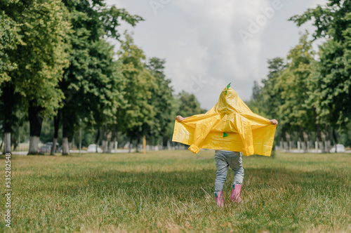 Happy little girl in yellow raincoat and muddy rubber boots running on dirt road through green grass near blooming rape seed field holding rhino in hand. Carefree childhood. photo