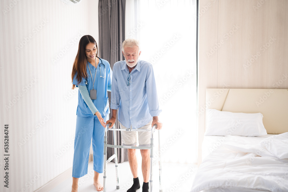 Portrait of Asian young nurse helping old elderly disable man grandfather to walk by using walker equipment in the bedroom. Senior patient of nursing home moving with walking frame and nurse support Portrait of Asian young nurse helping old elderly disable man grandfather to walk by using walker equipment in the bedroom. Senior patient of nursing home moving with walking frame and nurse support