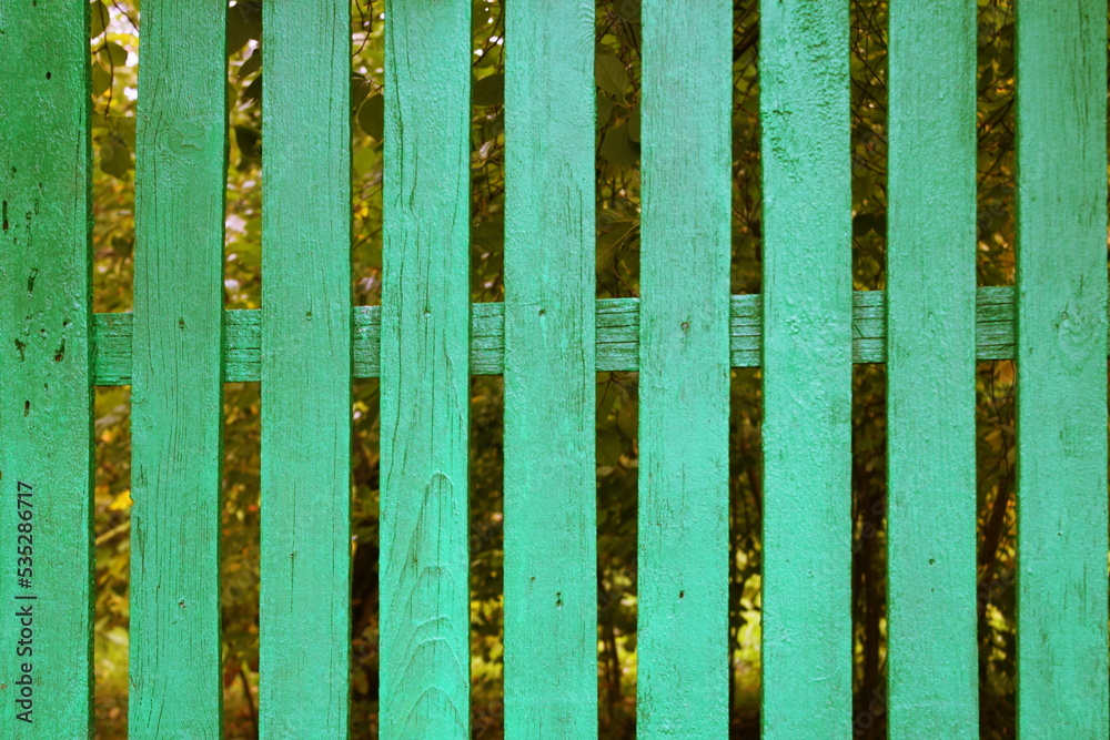 Green old fence. A garden with fruit trees is visible behind the fence. Green old fence. A garden with fruit trees is visible behind the fence.