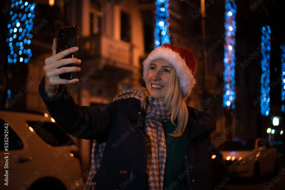Happy young woman in a Santa Claus hat taking a selfie against the background of the Christmas lights of the night city. Concept of holidays and people Happy young woman in a Santa Claus hat taking a selfie against the background of the Christmas lights of the night city. Concept of holidays and people
