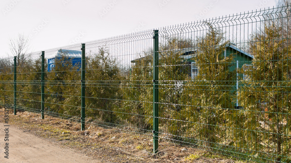 a fence made of 3d mesh, in the photo a fence against a background of green trees and gray sky a fence made of 3d mesh, in the photo a fence against a background of green trees and gray sky