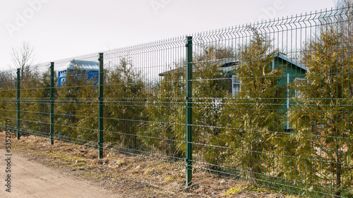 a fence made of 3d mesh, in the photo a fence against a background of green trees and gray sky © fotofotofoto