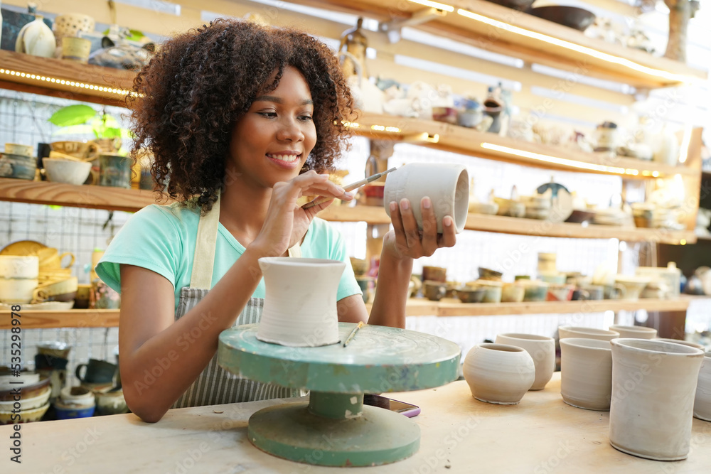 Close-up of concentrated beautiful craftswoman in apron sitting at pottery wheel and using craft tool while shaping wet clay vessel Close-up of concentrated beautiful craftswoman in apron sitting at pottery wheel and using craft tool while shaping wet clay vessel