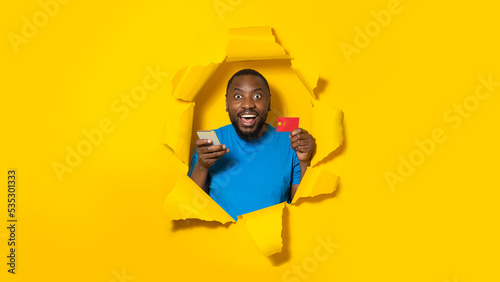 Excited black man holding credit card, using cellphone for online shopping, breaking through yellow paper background © Prostock-studio