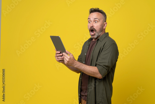 Portrait of impressed positive middle-age man holding in hands tablet open mouth isolated on yellow color background. Astonished bearded man received message