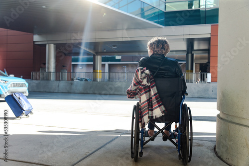 Young woman living with a disability in a wheelchair on the sidewalk at the airport waiting for an Uber to pull up on the road while traveling