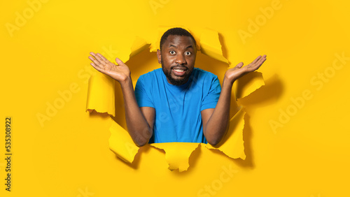 African american man spreading hands while complaining  posing in torn yellow paper hole
