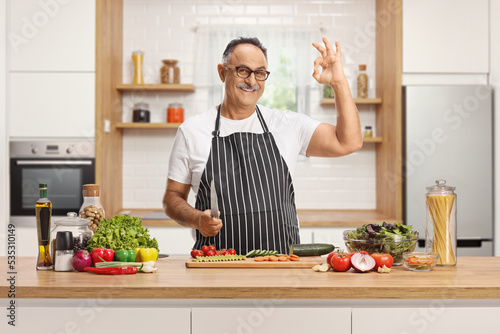 Mature man behind a kitchen counter holding a knife and gesturing ok
