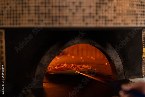 Pizza concept. Preparing traditional italian pizza. Long shovel for pizza, baking dough in a professional oven with open fire in interior of modern restaurant kitchen © arthurhidden