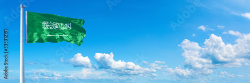 Saudi Arabia flag waving on a blue sky in beautiful clouds - Horizontal banner photo