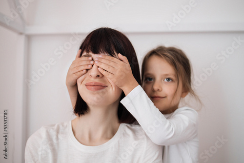 Satisfied caucasian preschooler girl closes eyes to young lady and congratulates with birthday, sitting on bed in bedroom interior. Surprise, holiday, love and relationship. Celebrate at home