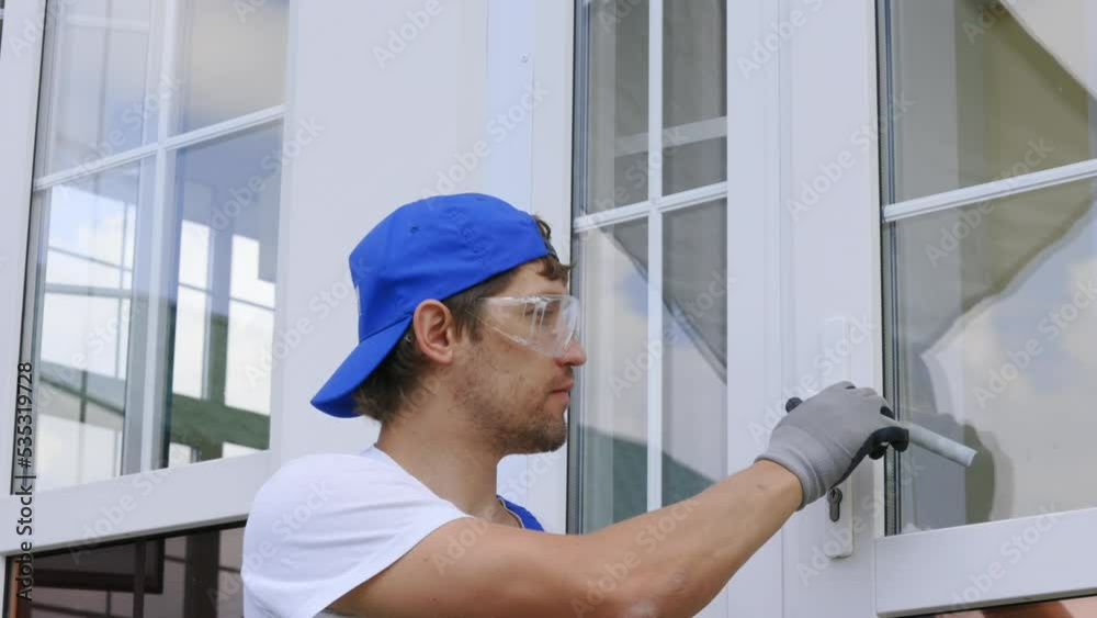 Worker in a blue cap and safety glasses with a screwdriver adjusts the door handle and lock on the white plastic door. Seasonal maintenance of plastic window locks by a professional craftsman Worker in a blue cap and safety glasses with a screwdriver adjusts the door handle and lock on the white plastic door. Seasonal maintenance of plastic window locks by a professional craftsman