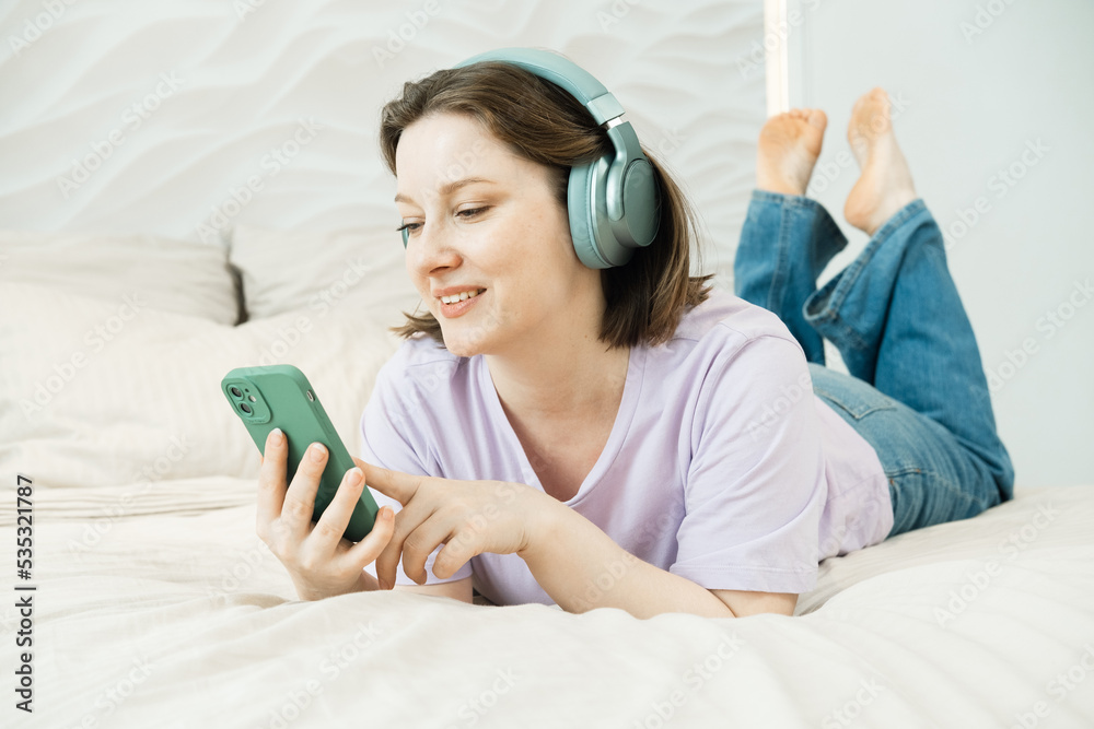 Smiling adult woman lies on the bed in headphones and listens to music Smiling adult woman lies on the bed in headphones and listens to music