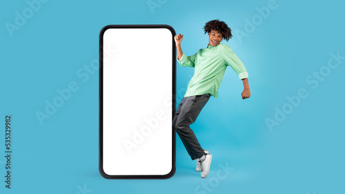 Cheerful black teenage guy standing on tiptoes near big blank smartphone