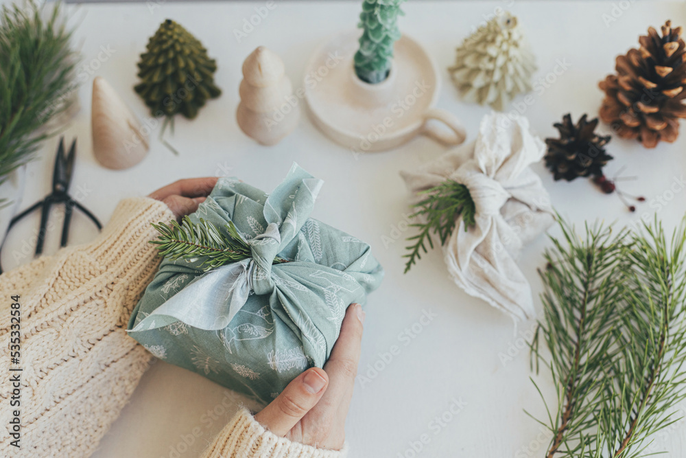 Zero waste Christmas. Hands holding gift box wrapped in green fabric with fir branch on white wooden table with eco friendly decor and candle. Furoshiki gift wrapping. Merry Christmas! Zero waste Christmas. Hands holding gift box wrapped in green fabric with fir branch on white wooden table with eco friendly decor and candle. Furoshiki gift wrapping. Merry Christmas!