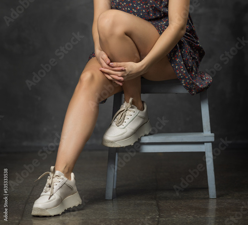 Beautiful female legs. The girl is sitting on a chair on a concrete floor, her legs are in white sneakers