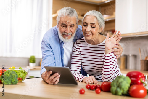 Video Call. Smiling Elderly Couple Using Digital Tablet In Kitchen For Teleconference