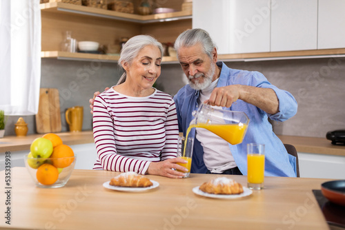 Elderly Spouses Eating Croissants And Drinking Juice While Having Breakfast In Kitchen