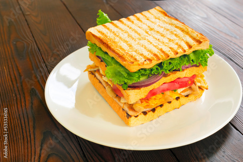 Homemade club sandwich with toasted bread slices, chicken breasts, tomato, salad leafs, cucumber, onion on white plate. On wooden tabletop. Side view.