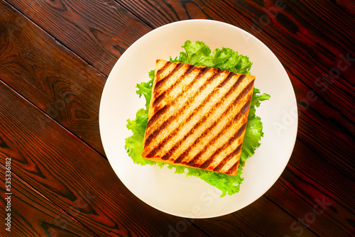 Homemade club sandwich with toasted bread slices, chicken breasts, tomato, salad leafs, cucumber, onion on white plate. On wooden tabletop. Flat lay. 