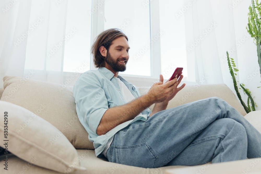 A man with a beard sits on the couch during the day and smiles at home and looks at his phone relaxing on his day off, life online on his phone texting with friends A man with a beard sits on the couch during the day and smiles at home and looks at his phone relaxing on his day off, life online on his phone texting with friends