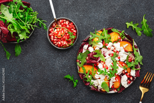 Fresh salad with arugula, radicchio, peaches, feta cheese and pomegranate seeds. Black table background, top view