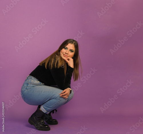 full length shot of a beautiful woman in makeup sitting on squat's position touching her chin and knee ahead a purple background