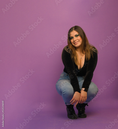full length image of a caucasian young lady sitting on squat position and touching her knees while looking at camera ahead a purple background