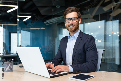 Portrait of mature boss inside office with laptop, successful and satisfied investor manager looking at camera and smiling man in glasses and business suit, investor with beard sitting on chair © Liubomir