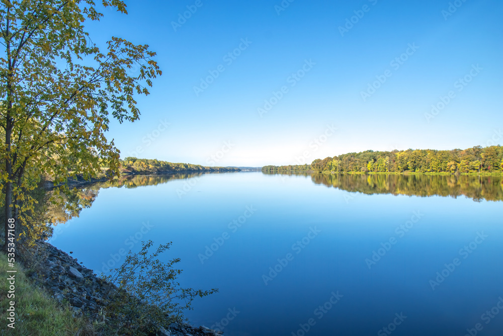View of a long, completely calm blue Ottawa River with forested banks reflected in water, rocky shore in foreground, blue sky, daytime, nobody