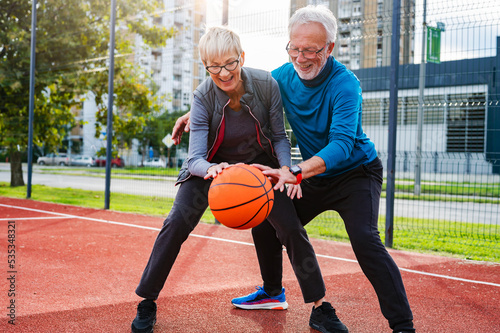 Cheerful active senior couple playing basketball on the urban basketball street court. Happy living after 60.