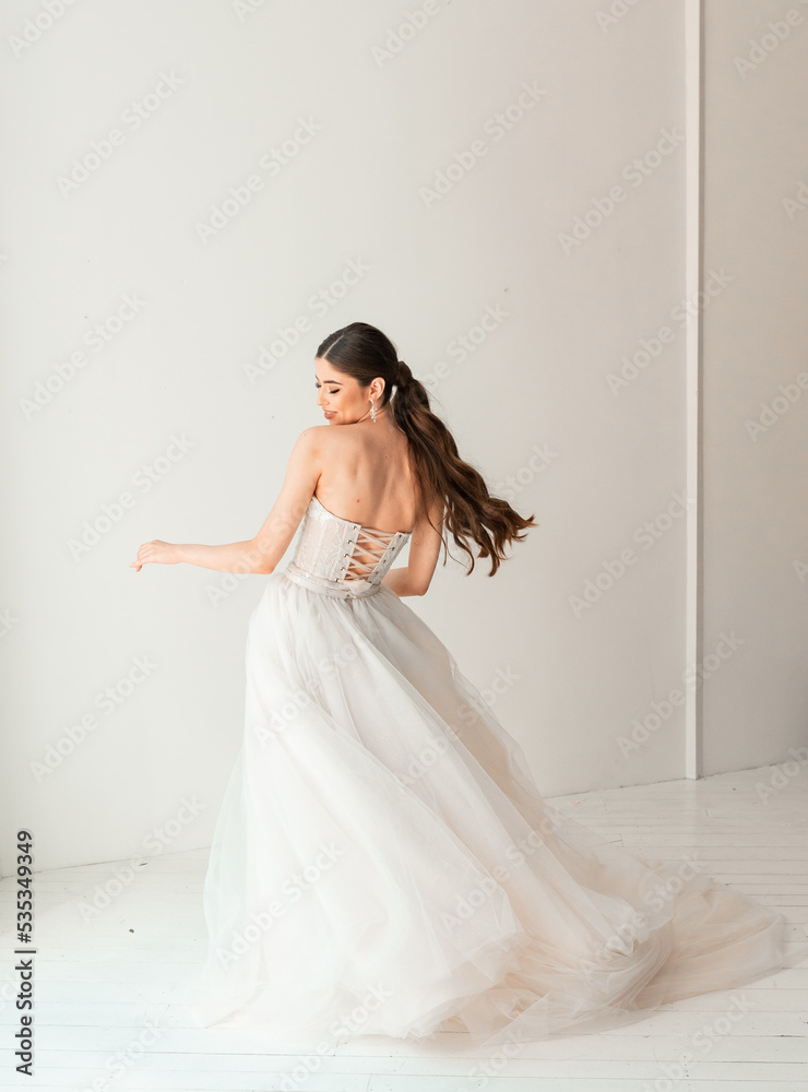 Elegant beautiful caucasian bride with long wavy brunette hair wearing white wedding dress and posing in bright interior Elegant beautiful caucasian bride with long wavy brunette hair wearing white wedding dress and posing in bright interior