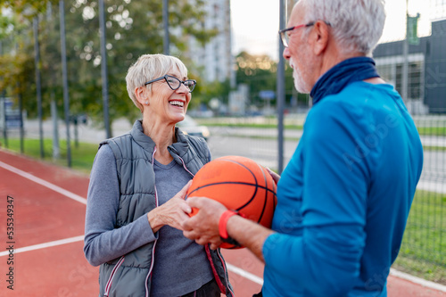 Portrait of cheerful active senior couple holding basketball ball standing on the urban basketball street court. Happy living after 60. © lordn