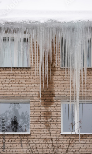 Large beautiful icicles hang from the snow-covered roof in front of the windows of the house. Snow on the roof. © goodmoments
