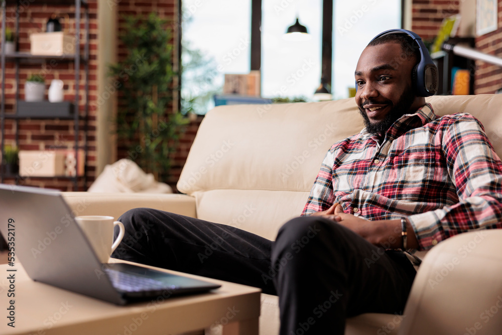 African american freelancer listening to music or podcast show on online website, using social media and headphones to work remotely. Working on laptop at home and enjoying song on internet at home.