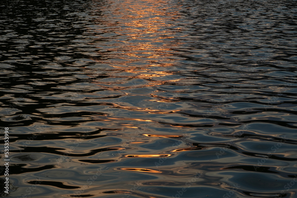 Dark blue water surface with ripples and golden sun light reflected on it at sunset. Natural background. Dark blue water surface with ripples and golden sun light reflected on it at sunset. Natural background.
