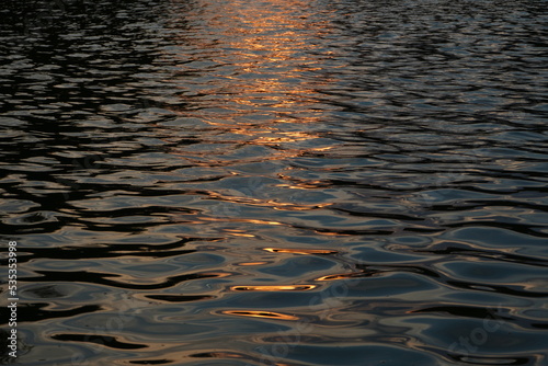 Dark blue water surface with ripples and golden sun light reflected on it at sunset. Natural background.