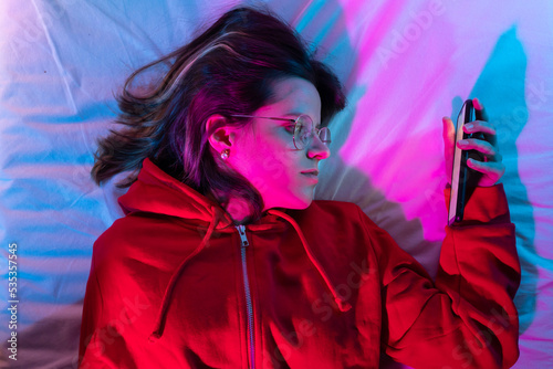Young girl looking her smart phone doom scrolling on bed in the middle of the night with neon lights. Technology at bed concept.